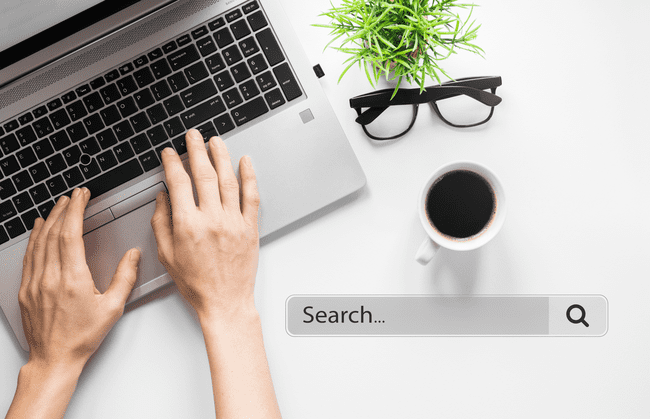 Hands typing on a laptop keyboard next to a coffee cup, glasses, and a small plant, with a search bar graphic overlay.