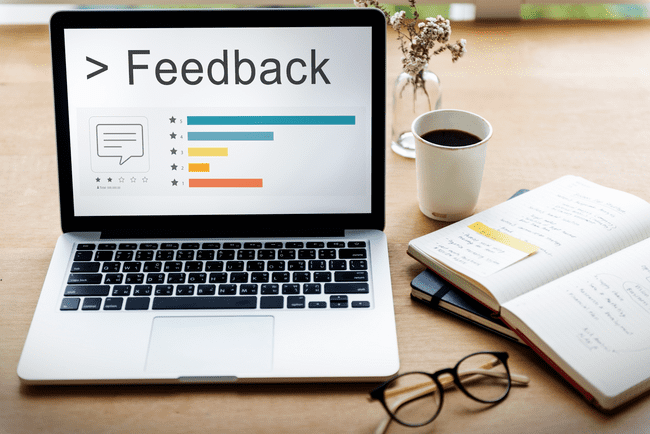 A laptop displaying a feedback screen with ratings and comments, next to a notebook, glasses, and a coffee cup on a wooden desk.