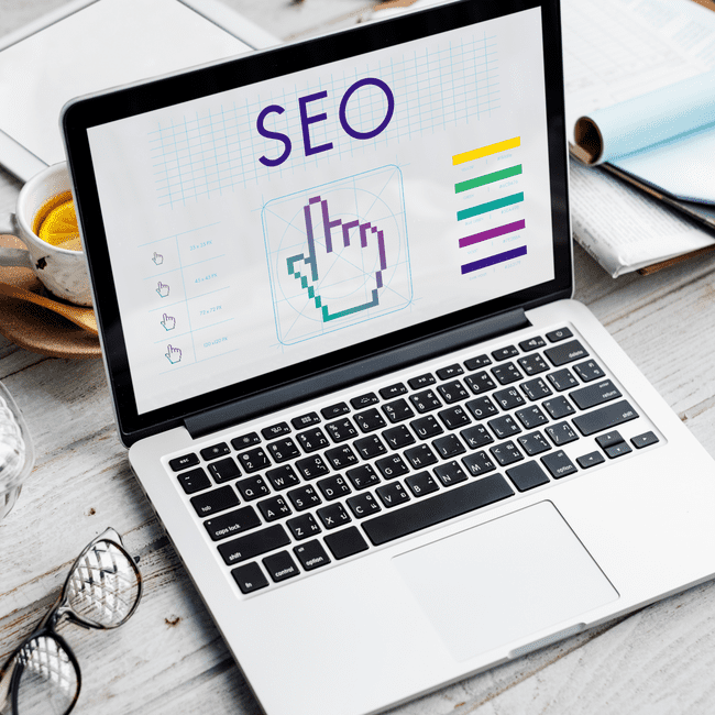 A laptop displaying the word "SEO" with various digital graphics and charts, placed on a desk with a cup of tea, glasses, and documents.
