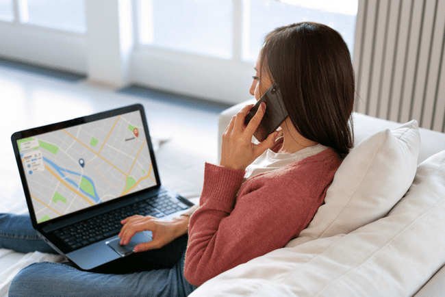 A woman sitting on a couch, talking on the phone while looking at a map on her laptop screen.