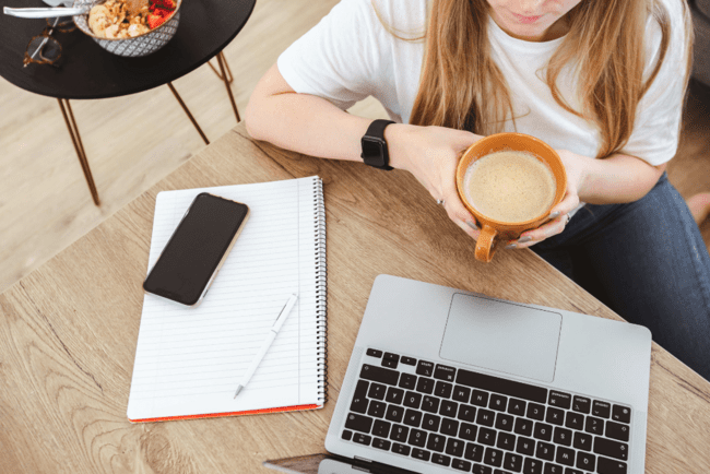 A business professional holding a cup of coffee, sitting at a desk with a laptop and notebook.
