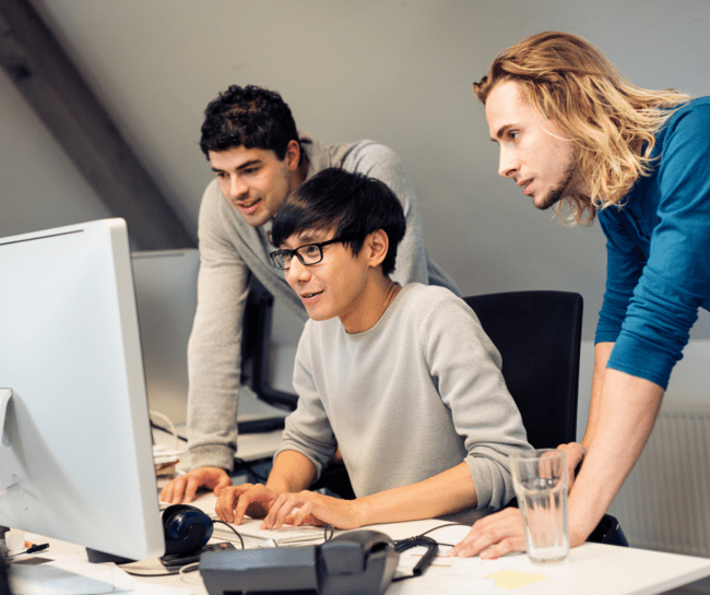 Three young professionals working together at a desk, looking at a computer screen.