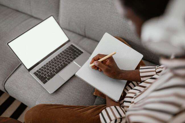 A person taking notes on a notepad while working on a laptop.