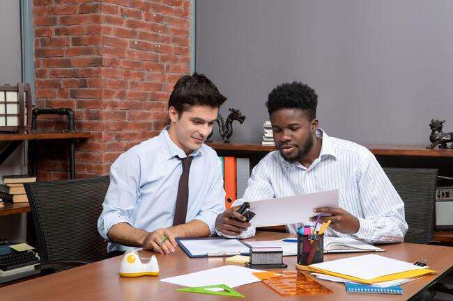 Two colleagues in a legal office setting, collaborating on documents and using a mobile phone for reference.