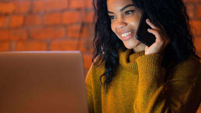A woman smiling while talking on the phone and working on a laptop.